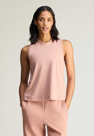 Kvinde med mellemtonet hud og skulderlangt mørkt hår iført en ærmeløs lyserød top og matchende bukser, hænderne i lommerne.