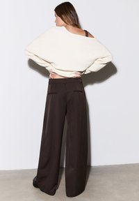 Pull en maille crème à épaules dénudées avec manches larges, associé à un pantalon large taille haute marron foncé avec poches arrière.