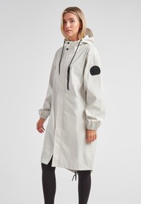 Cappotto impermeabile grigio chiaro con cappuccio, polsini elasticizzati e una patch nera sulla manica. Presenta una zip frontale e tasche.