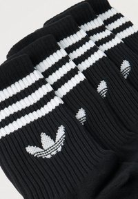 Juodų, gofruotų kojinių krūva, papuošta baltais Adidas trefoil logotipais ir dviem baltais horizontaliomis juostelėmis prie viršaus.
