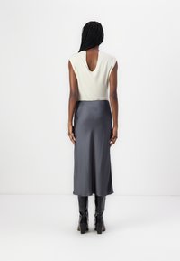 Kobieta z warkoczami stoi przodem do białej ściany, ubrana w beżowy top bez rękawów, szary midi spódnicę i czarne botki na obcasie.