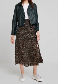 Mørkegrønt læder bikerjakke i kort model over en sort midi-nederdel med blomsterprint og sidelæg, parret med lysegrå sneakers.