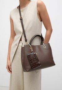 Braune Ledertasche mit strukturierter Form, ausgestattet mit einem abnehmbaren, strukturierten Geldbeutel, goldfarbenen Beschlägen und einem langen, verstellbaren Trageriemen.