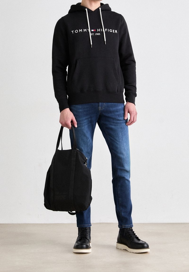 Person iförd en svart Tommy Hilfiger-hoodie, blå jeans, svarta stövlar, som håller en svart tote-väska mot en ljus, enfärgad bakgrund.