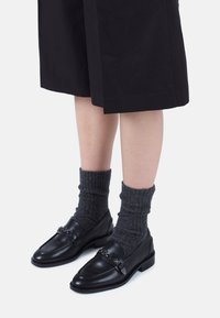Mocasines de cuero negro con punta redondeada y hardware decorativo, combinados con calcetines de canalé oscuro gris texturizado. Llevados con pantalones cortos holgados negros.