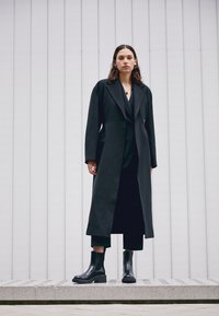 Svart lång kappa med en strukturerad design, känd med svarta ankelboots med en chunky sula; slät textur och skräddarsydd passform.