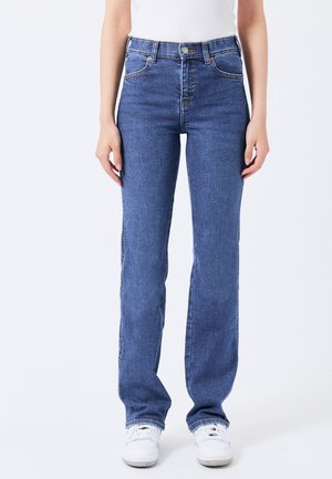 Osoba v modrých džínech s vysokým pasem a rovnými nohavicemi a v bílých teniskách stojí před jednoduchým bílým pozadím.