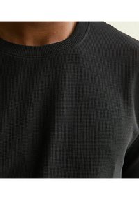 Czarny sweter o fakturowanej strukturze z prążkowanym wzorem, okrągłym dekoltem i dopasowanym kroju, prezentujący zbliżenie na powierzchnię materiału.