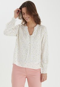Vit blus med små gula och rosa blommönster, knappslå med knappar, långa ärmar med volangkant. Bärs med rosa byxor.