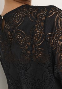 Czarna koronkowa bluzka z misternymi kwiatowymi wzorami, przezroczystą fakturą i wycięciem na plecach zapinanym na mały guzik.