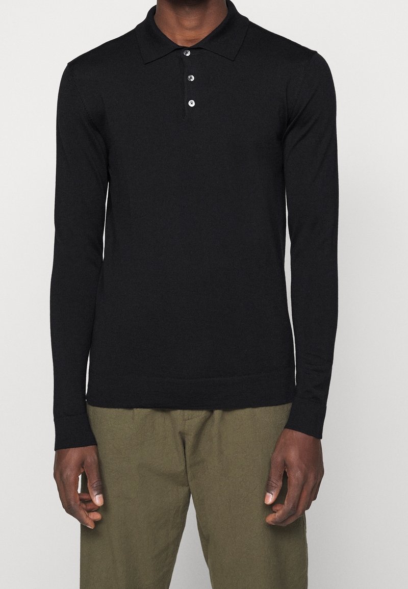 Polo noir à manches longues avec trois boutons, porté par une personne debout avec un pantalon vert olive sur un fond uni clair.