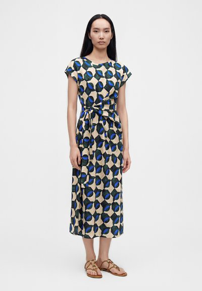 Femme portant une robe midi beige avec des motifs géométriques verts et bleus, des manches courtes, une taille nouée et des sandales plates marron.