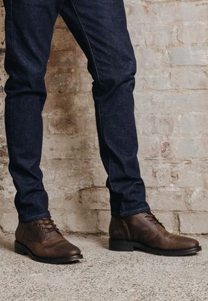 Braune Leder-Ankle-Boots mit Schnürdesign, runder Spitze und dunkler Sohle, kombiniert mit dunkelblauen Jeans vor einer strukturierten Wand.