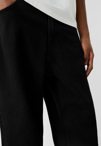 Svarta jeans i denim med lös passform. Närbild visar textur och sömnadsdetaljer, tillsammans med en vit t-shirt som delvis syns ovanför.