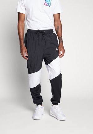 Homme portant un pantalon de survêtement Jordan noir et blanc, des baskets Air Jordan blanches et un t-shirt blanc avec un logo Jordan coloré.
