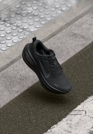 Chaussure de course Nike noire avec un swoosh réfléchissant flottant au-dessus d’un trottoir gris près d’un bord de trottoir texturé et d’une ligne blanche.