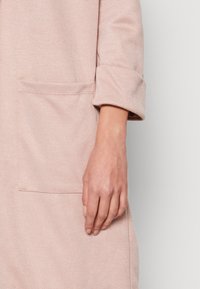 Vaaleanpunainen teksturoitu kangastakki, jossa on käärityt hihat ja tasku. Lähikuva kädestä, joka lepää taskun lähellä.