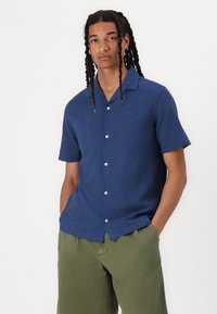 Short-sleeve navy blue linen shirt with a collar, featuring a button-down front and subtle texture. Worn with light green shorts.