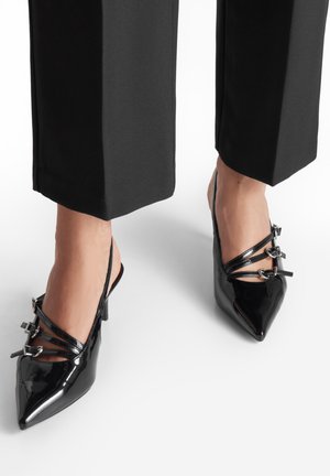 Nohy ve černých lodičkách na podpatku z lakované kůže se špičatou špičkou a třemi přezkami, doplněné černými širokými kalhotami se zkrácenou délkou.