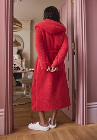 Red, fluffy bathrobe with a hood and tie waist, accompanied by white, soft slippers with floral embroidery, seen from the back.