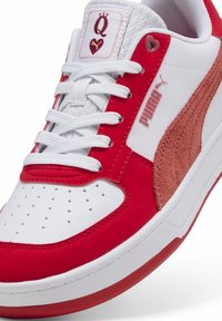 Baskets rouges et blanches avec une tige en cuir lisse, des accents en daim, des perforations sur le bout et une étiquette de languette texturée avec un logo en forme de cœur.