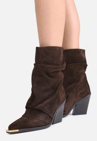 Botine din suede maro cu un design relaxat, vârf ascuțit și un accent aurit pe vârful pantofului. Au un toc pătrat pentru sustinere.