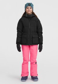 Černá péřová bunda se dvěma předními kapsami, doplněná jasně růžovými snowboardovými kalhotami. Tmavě modré boty a tyrkysová čepice dotvářejí celkový vzhled.