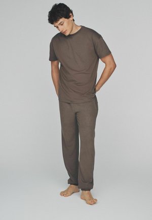 Junger Mann barfuß, schaut nach unten, Hände in den Taschen, trägt ein passendes braunes Kurzarmhemd und Hose vor einem schlichten Hintergrund.