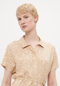 Beige shirt dress with short sleeves, featuring an all-over shell pattern in a lighter shade. Designed with a collar and button closure.
