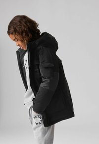 Chaqueta acolchada negra con capucha, con cierre frontal, bolsillos laterales y tejido texturizado. Lleva puesta una sudadera gris y pantalones.