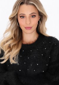 Donna bionda con capelli ondulati, che indossa orecchini a cerchio e un maglione nero di peluche decorato con piccole strass, che sorride dolcemente.