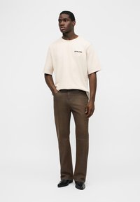 Bărbat stând cu mâinile în buzunare, purtând un tricou bej supradimensionat și pantaloni maro largi, împreună cu pantofi negri pe fundal alb.