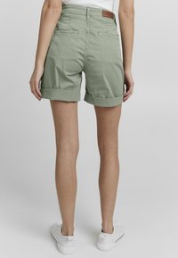 Shorts en coton vert avec des revers, dotés de poches arrière et d'une étiquette en cuir au niveau de la taille. Portés avec des baskets blanches.