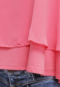 Blouse rose à couches avec un tissu doux et léger. L'ourlet présente un léger volant, assorti à la ceinture visible d'un jean en denim foncé en dessous.