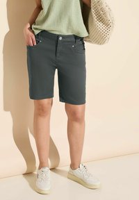 Pantaloni corti grigi in tessuto liscio, con due tasche frontali e un design aderente. Abbinati a una maglia verde a trama e sneakers bianche.