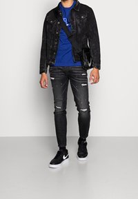 Persona vistiendo una chaqueta de mezclilla negra, camiseta gráfica azul, jeans negros desgastados, zapatillas Nike negras y una bolsa cruzada negra frente a un fondo sencillo.