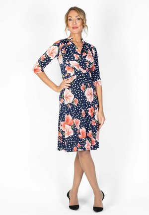 Frau steht mit einer Hand in der Hüfte, trägt ein marineblaues knielanges Kleid mit weißen Punkten und großem rosa Blumenmuster, schwarze High Heels.