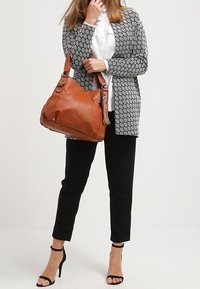 Bruine leren schoudertas met twee schouderbanden, gestructureerde afwerking en decoratieve franjes. Draagt samen met een zwart-wit gebreide cardigan.