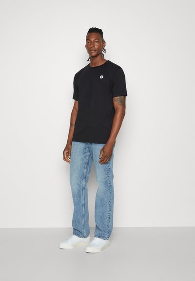 Svart bomull T-skjorte med rund hals og liten hvit logo, kombinert med lyseblå jeans og hvite joggesko. Minimalistisk design, uformell passform.