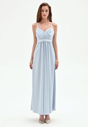 Donna che indossa un abito azzurro chiaro senza maniche con corpetto plissettato e fascia in vita, in piedi dritta contro uno sfondo bianco semplice.