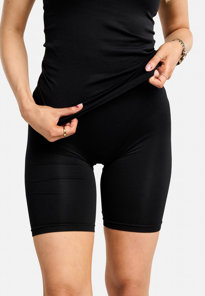 Personne portant un short et un haut noirs ajustés, tirant sur le t-shirt pour montrer l'extensibilité et la coupe du tissu sur fond blanc.
