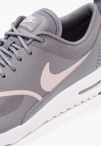 Detailní záběr na šedou sneaker Nike Air Max Thea s r�ůžovým logem swoosh a bílou podrážkou na bílém pozadí.