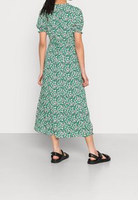 Zelené květinové midi šaty s nabíranými rukávy, s úzkým pasem a rozptýlenými bílými a růžovými květy. Nosené s černými sandály.