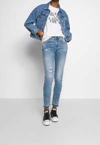 Chaqueta de mezclilla en azul claro, camiseta blanca con texto impreso, jeans skinny desgastados en azul claro y zapatillas negras con acentos blancos.