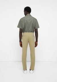 Camisa de manga corta verde oliva con un patrón sutil, combinada con pantalones beige ajustados y zapatillas blancas. Vista trasera que muestra los detalles del diseño.