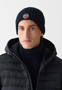 Căciulă tricotată de culoare bleumarin cu un model reliefat de nervuri și o patch cu logo rotund. Asortată cu o geacă puf vărgată neagră și un pulover închis cu guler înalt.