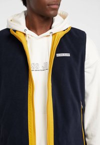 Marineblaues Fleeceweste mit gelbem Reißverschluss und Applikationen. Verfügt über ein Logoabzeichen und wird über einem weißen Kapuzenpullover mit aufgedrucktem Text getragen.