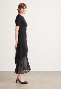 Abito nero con maniche corte, busto aderente, gonna semi-trasparente e lunghezza sotto il ginocchio. Abbinato a tacchi slingback neri.