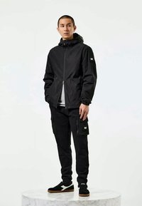 Jaunas vyras stovi ant apvalaus marmurinio pjedestalo, apsirengęs juodu džinsiniu kailiniu su gobtuvu, juodomis krovinių kelnėmis ir juodais sportiniais bateliais su baltais akcentais.