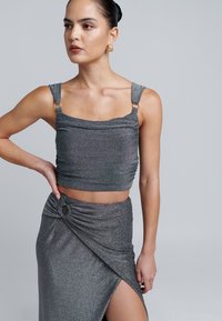 Błyszczący szary top w fasonie crop z marszczeniem i regulowanymi ramiączkami. Pasująca spódnica z wysokim rozcięciem, ozdobiona skręconym węzłem i okrągłym akcentem.